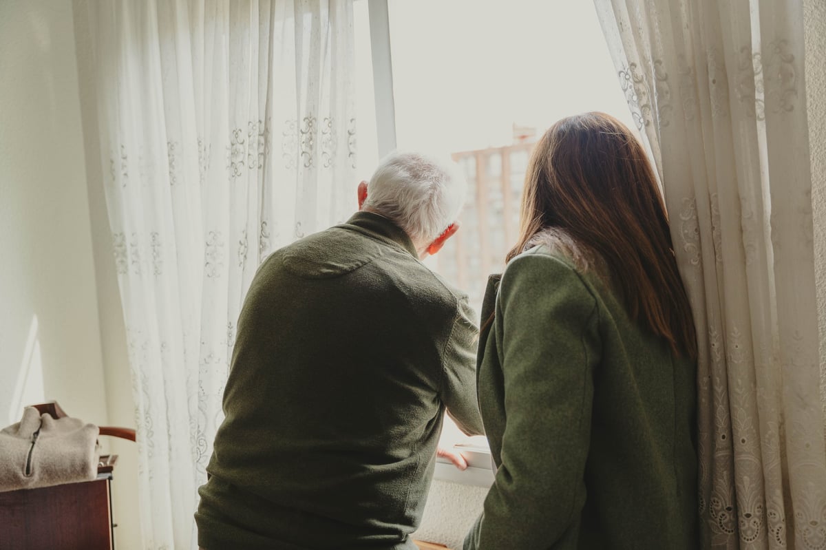 Een oudere mevrouw kijkt met een familielid naar buiten vanaf de bank in de woonkamer.
